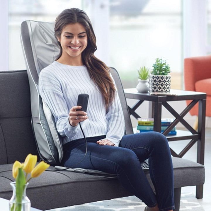 Woman sits in chair and looks at controller while enjoying a massage from the Homedics Gentle Touch Gel Deluxe Shiatsu Massage Cushion with Soothing Heat