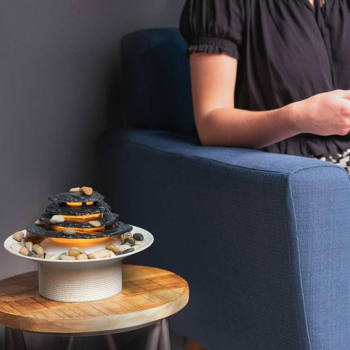 Woman sits in chair next to Homedics Mirra Zen Tabletop Relaxation Fountain on table