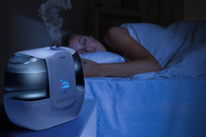 A woman sleeps soundly in her bed while the TotalComfort® Ultrasonic Humidifier with UV-C Technology and Remote Control sits on her nightstand.