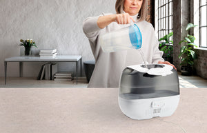 A woman pours water from the top of the TotalComfort® Ultrasonic Humidifier with UV-C Technology and Remote Control