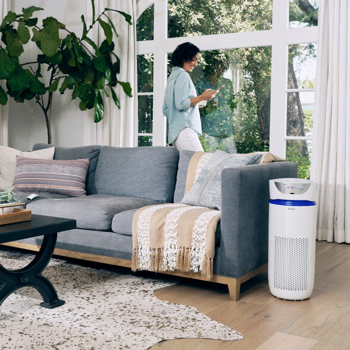 White | Homedics TotalClean 5-in-1 UV-C Deluxe Large Room Air Purifier sits next to couch and purifies air while woman stands and checks messages on phone by the window
