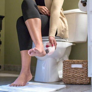 Woman in sits in bathroom while holding the Homedics SoftSilk Rechargeable Pedicure Callus Remover next to foot