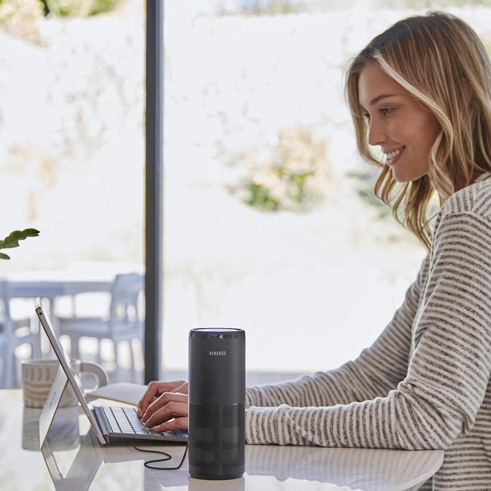 Black | Woman smiling while typing on tablet next to the Homedics TotalClean 4-IN-1 UV Portable Air Purifier in black