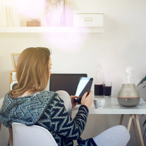 Steam is coming out of Homedics® diffuser and a woman inhales the same for calm experience while engaging herself in mobile.