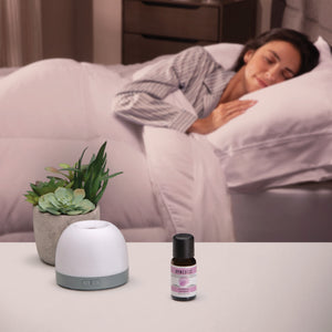 Aromatherapy diffuser and Homedics Lavender Essential Oil 15 ml sits on table with woman sleeping in bed in the background