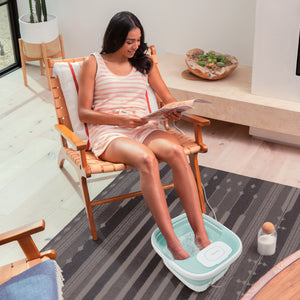 A woman uses the Homedics® Smart Space Elite Footbath with Heat Boost as she sits in a chair and reads a magazine.