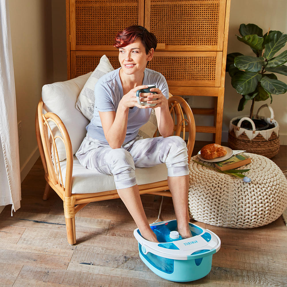 Smiling woman sits in chair and holds mug with feet in the Homedics Shower Bliss Foot Spa