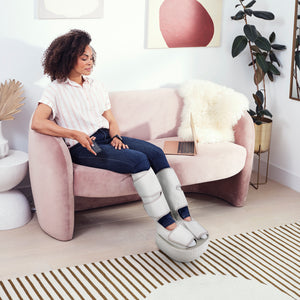 Woman in blue jeans sits on pink couch while wearing the Homedics Real Relief Leg & Foot Massager
