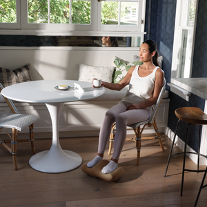 Woman enjoying her morning coffee sitting at a table while massaging her feet with the Homedics Comfy Footrest Massager
