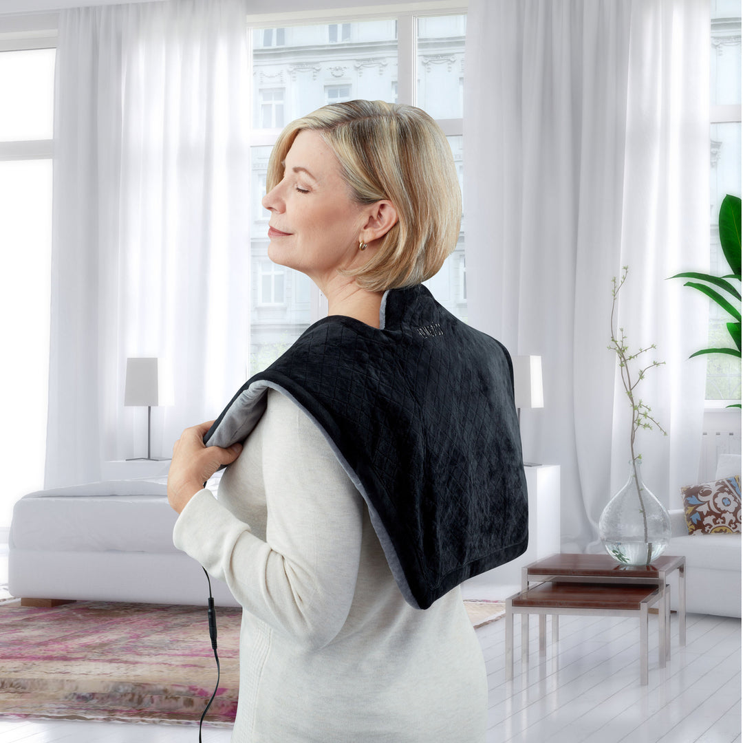 A woman wears the Homedics Thera-P Heated Neck and Shoulder Massager around her shoulders and enjoys the muscle relief