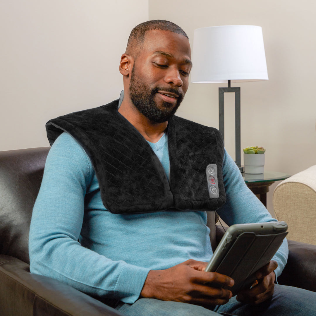 A man wears the Homedics Thera-P Heated Neck and Shoulder Massager while relaxing on the couch reading an electronic book