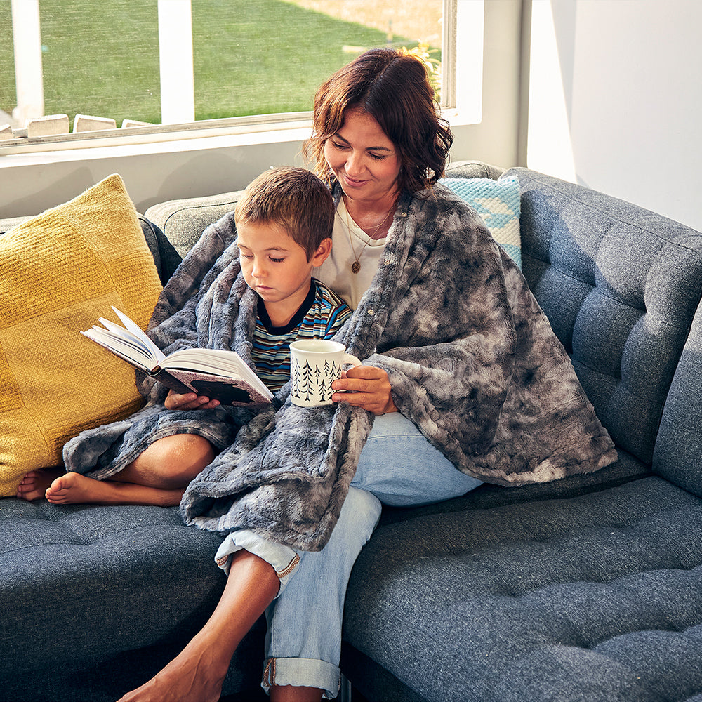 Woman reads book on couch with child while wrapped in the Homedics Cordless Throw & Cape with Soothing Heat & Vibrating Massage