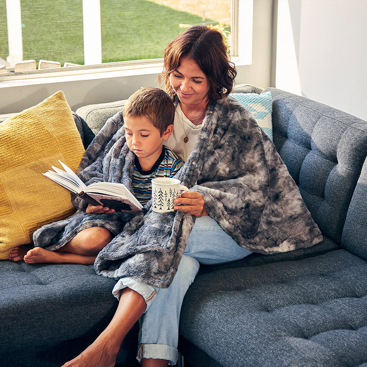 Woman reads book on couch with child while wrapped in the Homedics Cordless Throw & Cape with Soothing Heat & Vibrating Massage