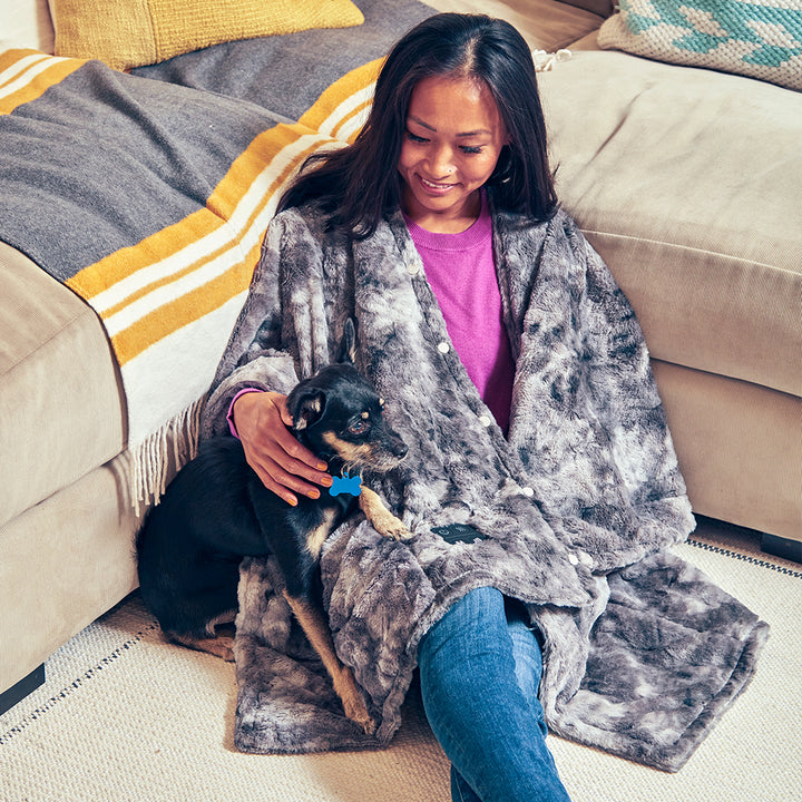 Woman sits on floor with dog next to sofa while wrapped in the Homedics Cordless Throw & Cape with Soothing Heat & Vibrating Massage