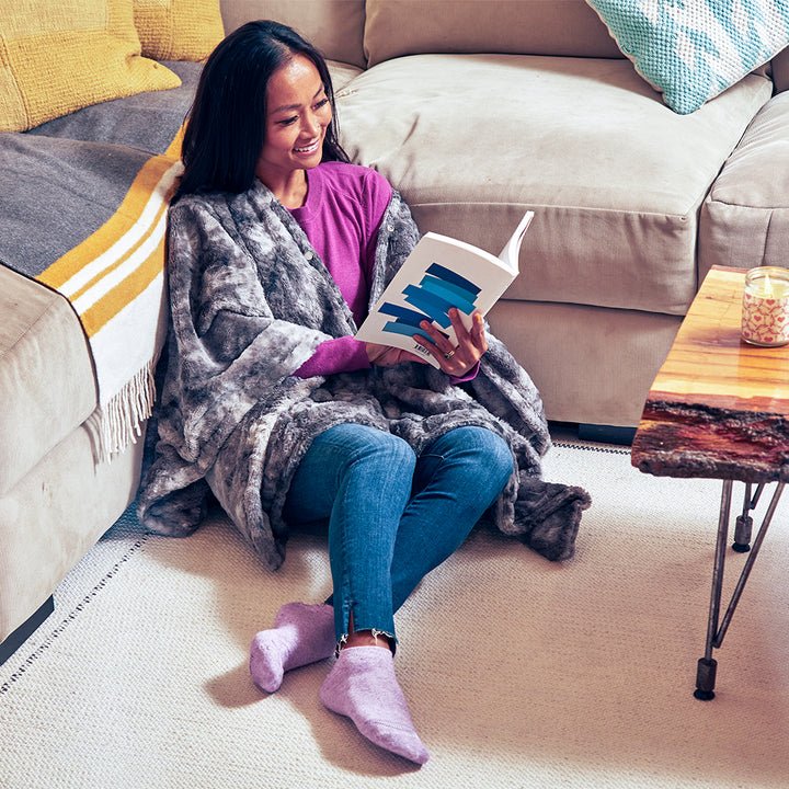 Woman sits on floor next to sofa reading book while wrapped in the Homedics Cordless Throw & Cape with Soothing Heat & Vibrating Massage