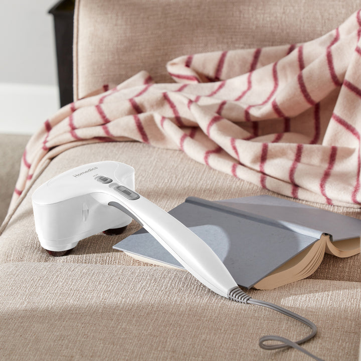 Homedics® Duo Percussion Body Massager with Heat lays on the couch next to an open book