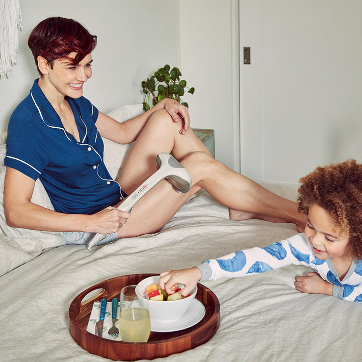 Brown | Woman sits on bed with small child while enjoying a leg massage with the Homedics Percussion Action Plus Handheld Massager with Heat