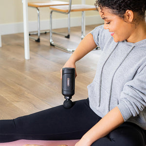 Smiling woman sits on floor while massaging her right thigh with the Homedics Therapist Select Pro Percussion Massager