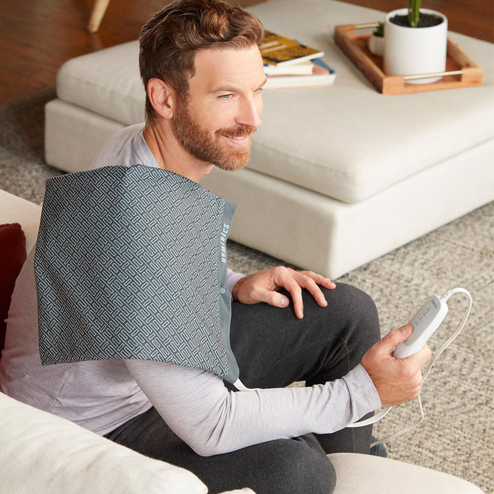 Back angle view of man smiling while sitting on couch with the Homedics 12" x 15" Heating Pad with Removable Cover draped around right shoulder