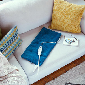 Angled view of the Homedics 12 x 24 Weighted Heating Pad with Humiditech on white couch with colorful pillows