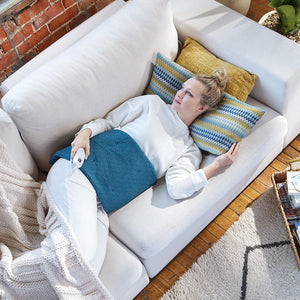 Woman lays on couch with abdomen covered by the Homedics 12 x 24 Weighted Heating Pad with Humiditech