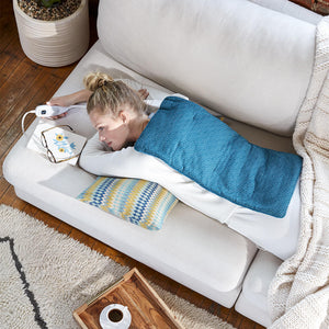 Woman lays on her stomach on couch with the Homedics 12 x 24 Weighted Heating Pad with Humiditech on her back