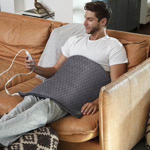 Man sits on couch looking at the LCD controller of the Homedics 24 x 24 Plush Oversized Heating Pad that is spread across his abdomen and lap