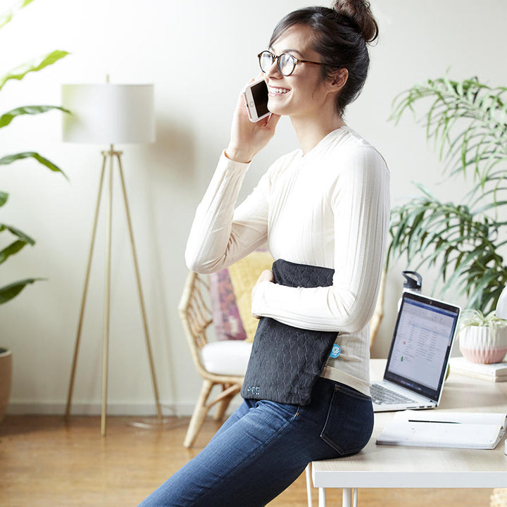 Woman sits on the edge of desk and talks on the phone while holding the Homedics 12" x 15" Weighted Gel Heating Pad against her stomach