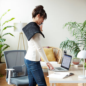 Woman standing in front of a laptop talks on the phone with  the Homedics Weighted Hot & Cold Gel Heating Pad resting on right shoulder