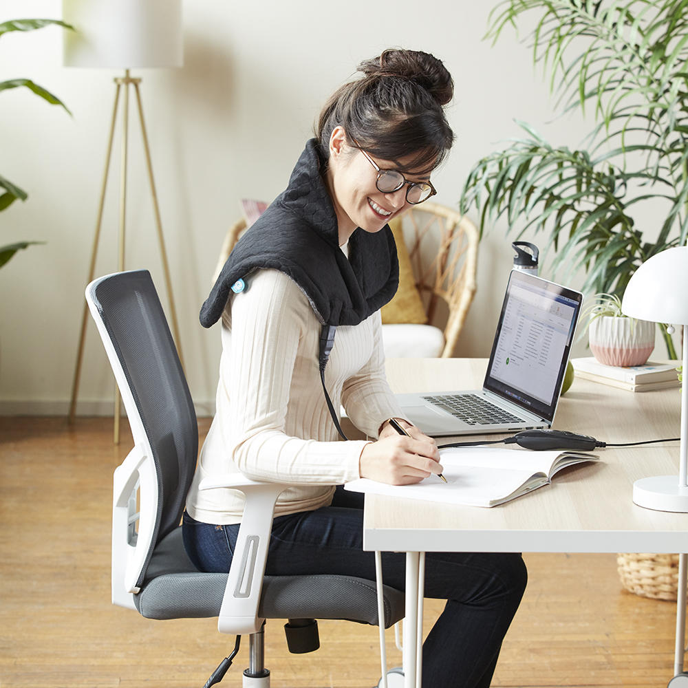 Woman wears the Homedics Weighted Gel Wrap on her shoulders while working at home office desk in brightly lit room