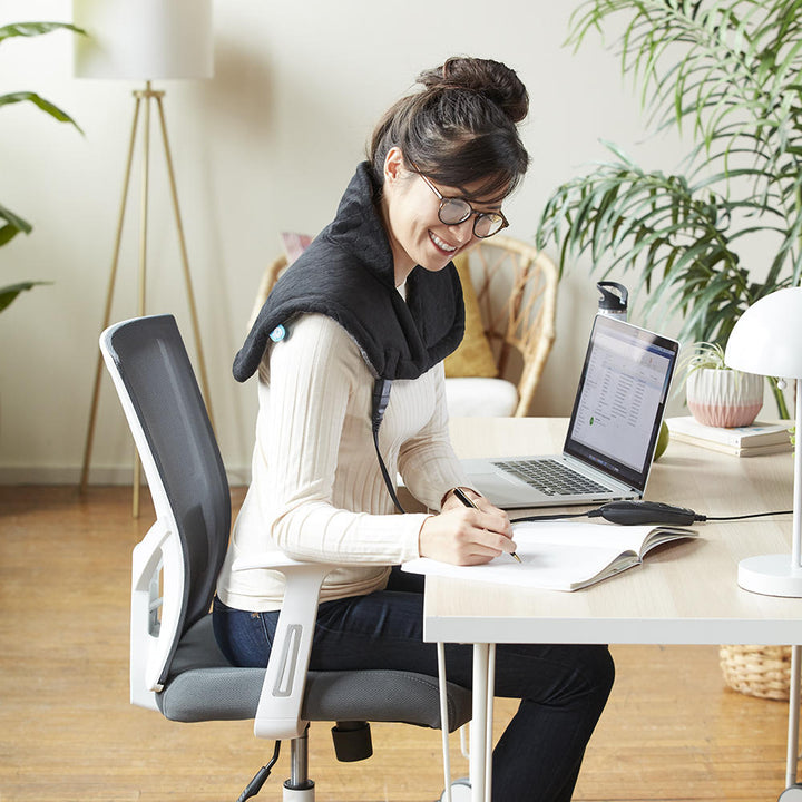 Woman wears the Homedics Weighted Gel Wrap on her shoulders while working at home office desk in brightly lit room