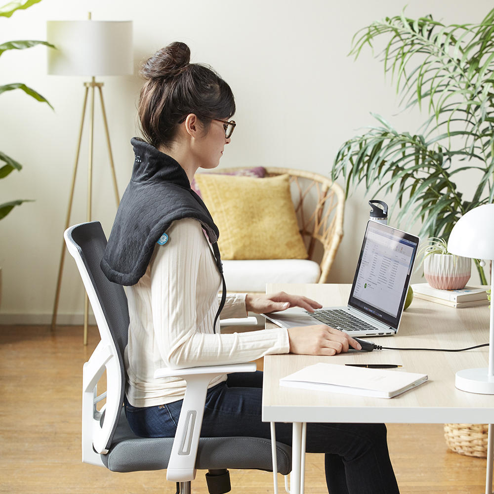 Alternate view of woman wearing the Homedics Weighted Gel Wrap on her shoulders while working at home office desk in brightly lit room