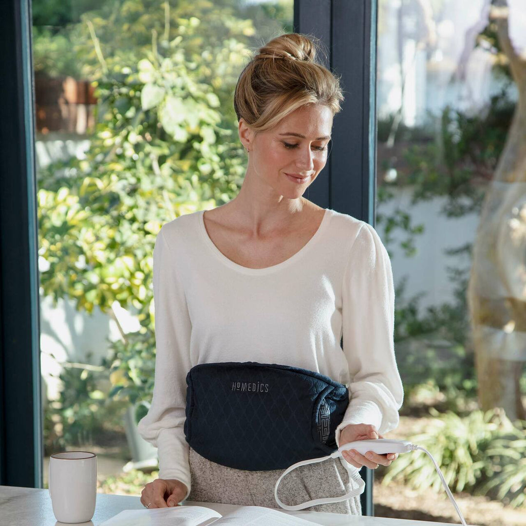 Woman standing at counter wearing the Homedics Women's Health Comfort Heating Belt uses controller to adjust the settings