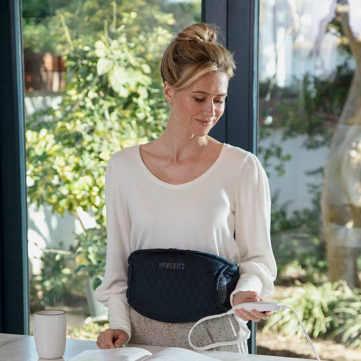 Woman standing at counter wearing the Homedics Women's Health Comfort Heating Belt uses controller to adjust the settings