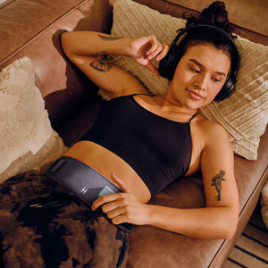 Homedics® Infrared + Red Light Pain Therapy Wrap is worn on the stomach to improve circulation while she reclines and listens to music on the couch