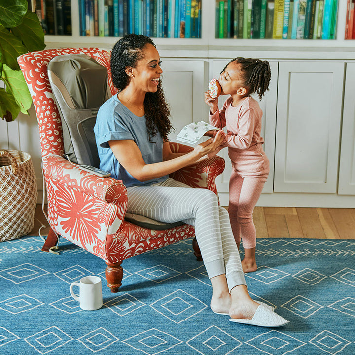 Woman sits in chair with small child while enjoying a massage from the Homedics Shiatsu Elite Pro Massage Cushion with Heat