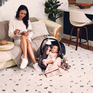 Woman relaxes on couch as baby in baby carrier reaches for the Homedics SoundSpa On-The-Go attached to carrier