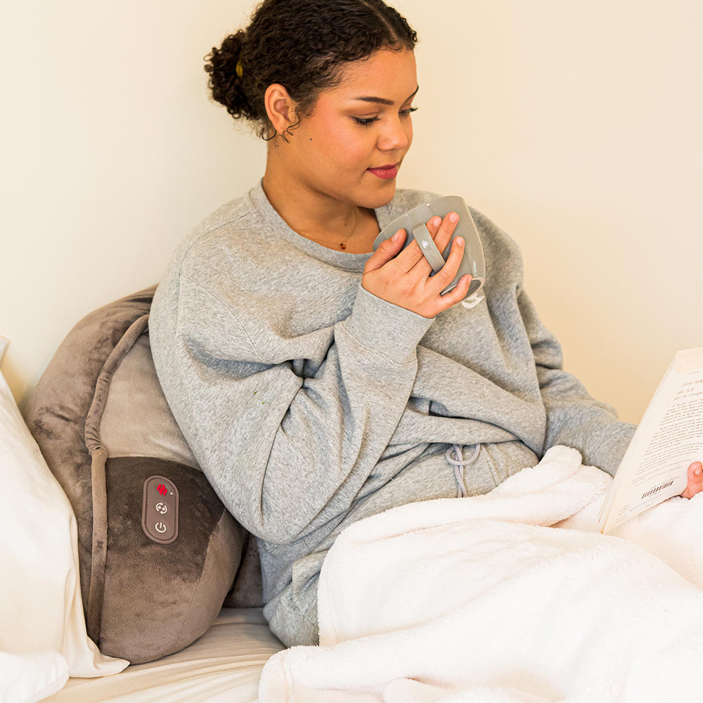 Woman lays in bed enjoying massage from the Homedics Shiatsu Bedrest Plus while reading book