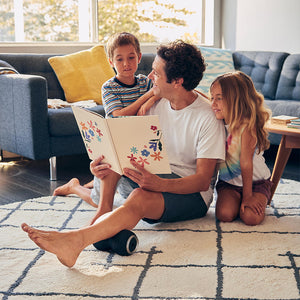 Man sits on floor and reads book to children while massaging left calf with the Homedics Double-Barrel Total Body Massager
