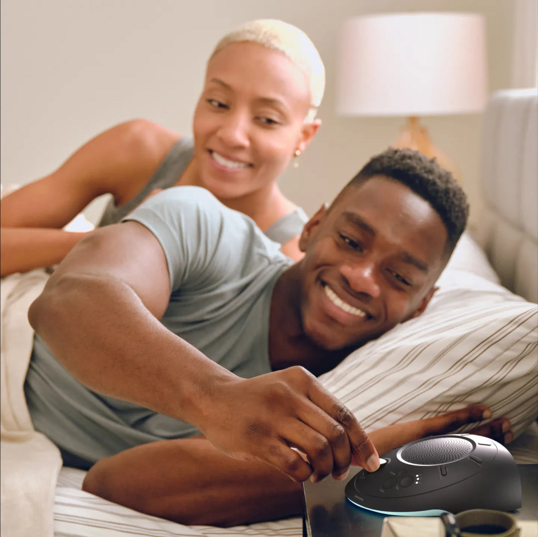 Night | Homedics Portable Sound Machine with Light placed on a bedside table, while a couple relaxes in bed