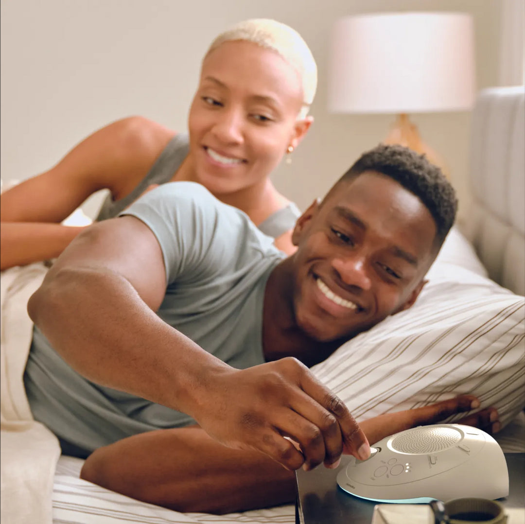 Dove | Homedics Portable Sound Machine with Light placed on a bedside table, while a couple relaxes in bed