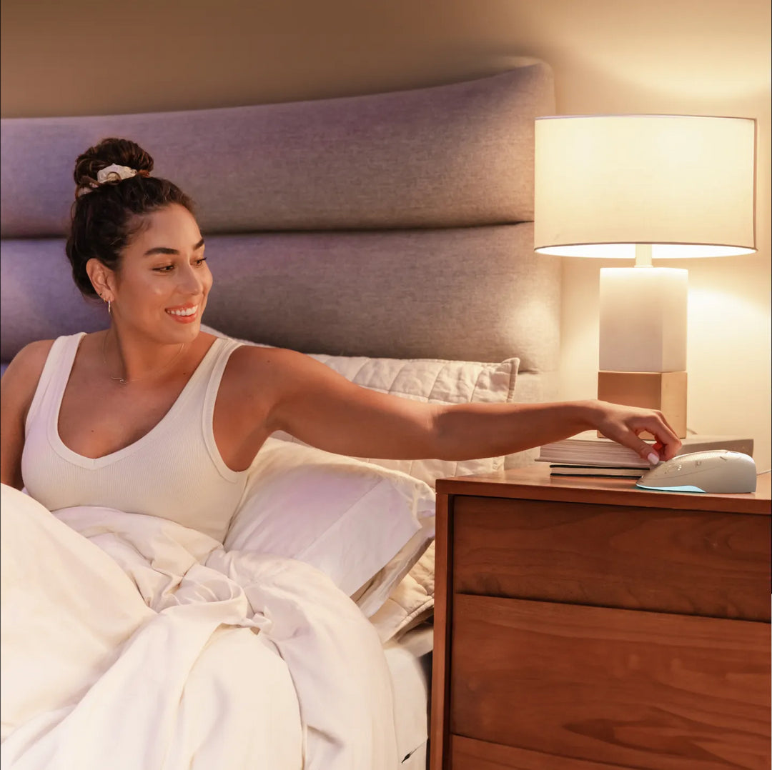 Dove | Homedics Portable Sound Machine with Light placed on a bedside table, while a woman relaxes in bed.