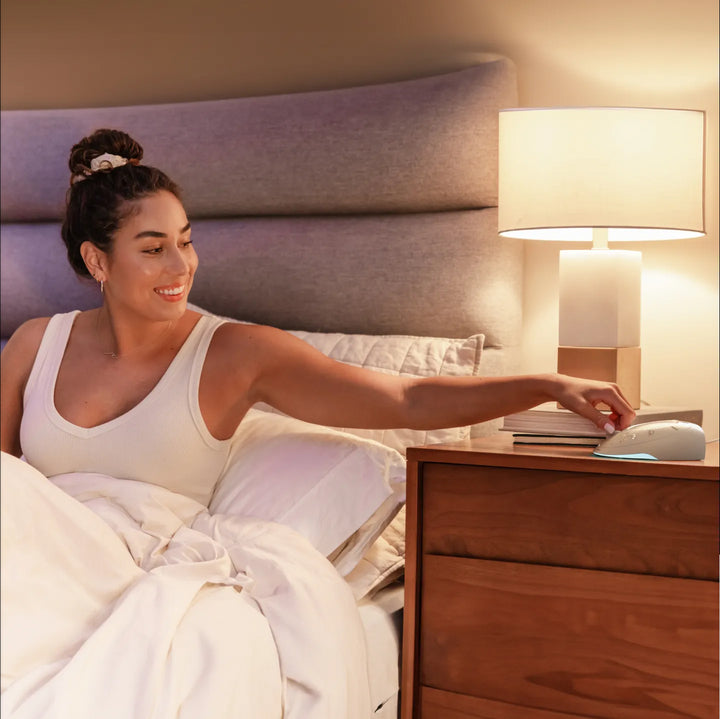 Dove | Homedics Portable Sound Machine with Light placed on a bedside table, while a woman relaxes in bed.