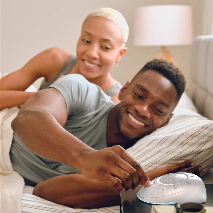 Cloud | Homedics Portable Sound Machine with Light placed on a bedside table, while a couple relaxes in bed