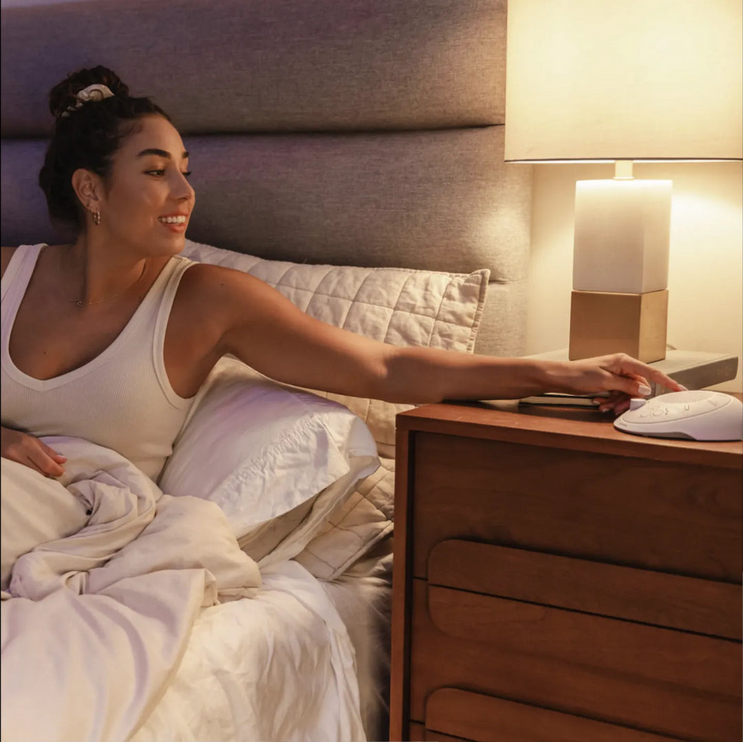 Cloud | Homedics Portable Sound Machine with Light placed on a bedside table, while a woman relaxes in bed.