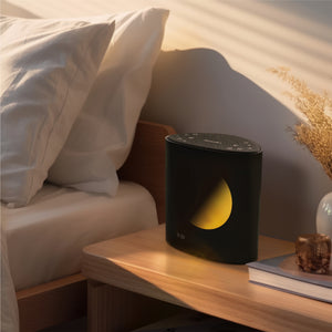 Lifestyle image of Sound Sleep Moon Dream Sound Machine & Clock placed on table besides bed.