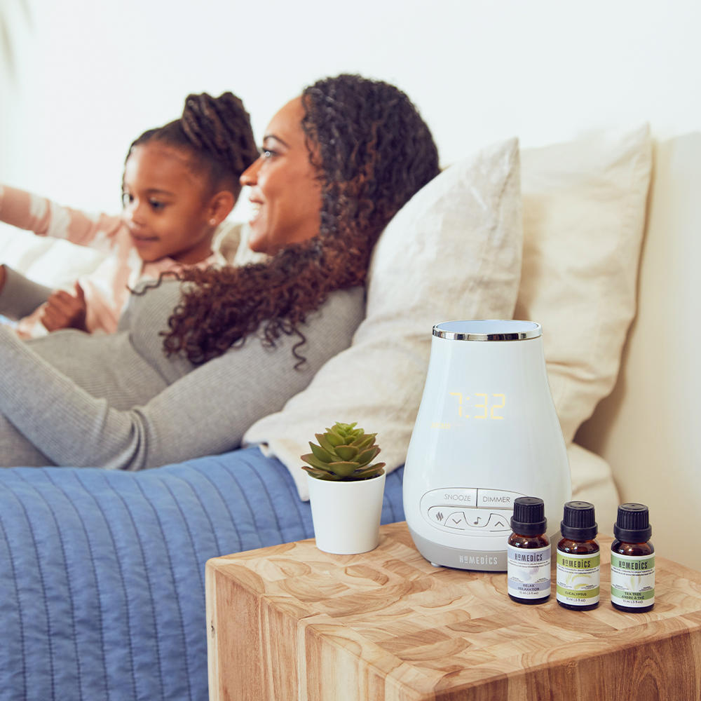 Woman lays in bed with child with the Homedics SoundSpa Slumber Scents on nightstand with essential oils
