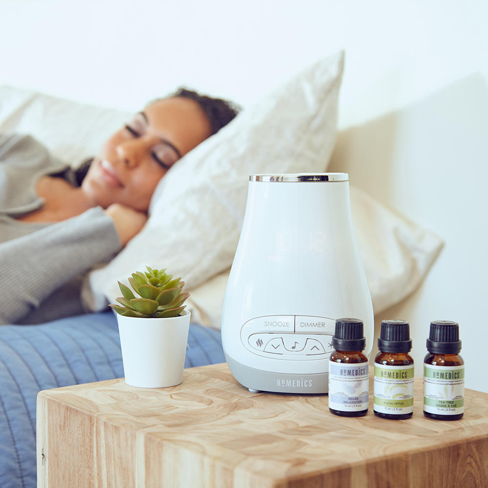 Woman sleeps in bed with the Homedics SoundSpa Slumber Scents on nightstand with essential oils