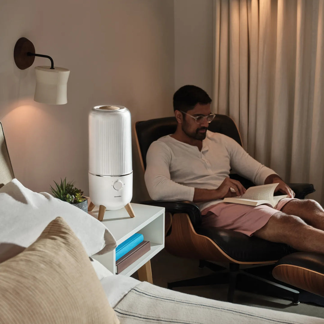 Man sitting in chair reading a book in his bedroom with the Homedics® Ultrasonic Humidifier on the table next to him