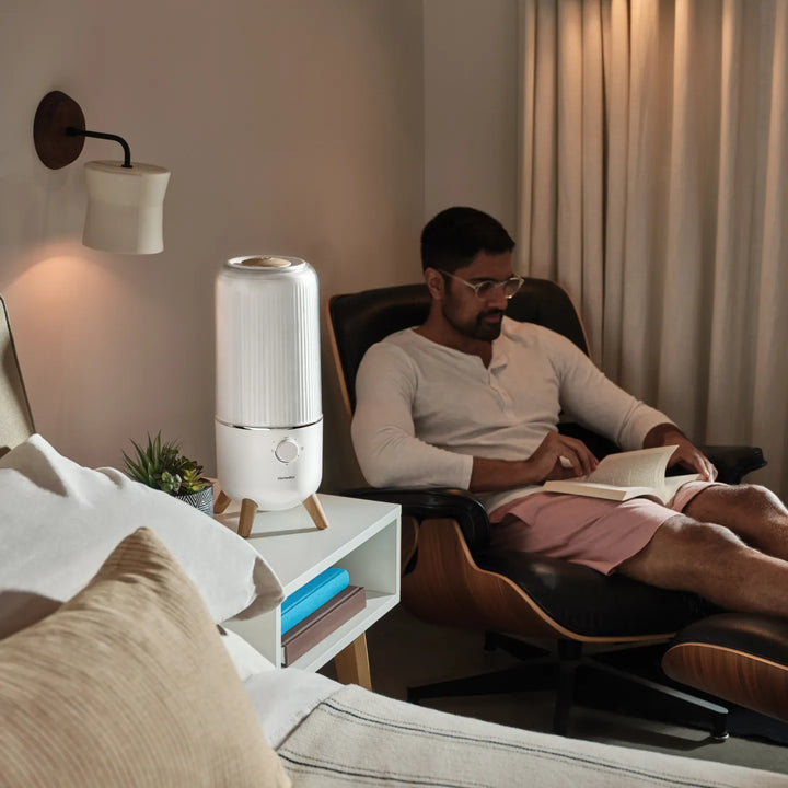 Man sitting in chair reading a book in his bedroom with the Homedics® Ultrasonic Humidifier on the table next to him